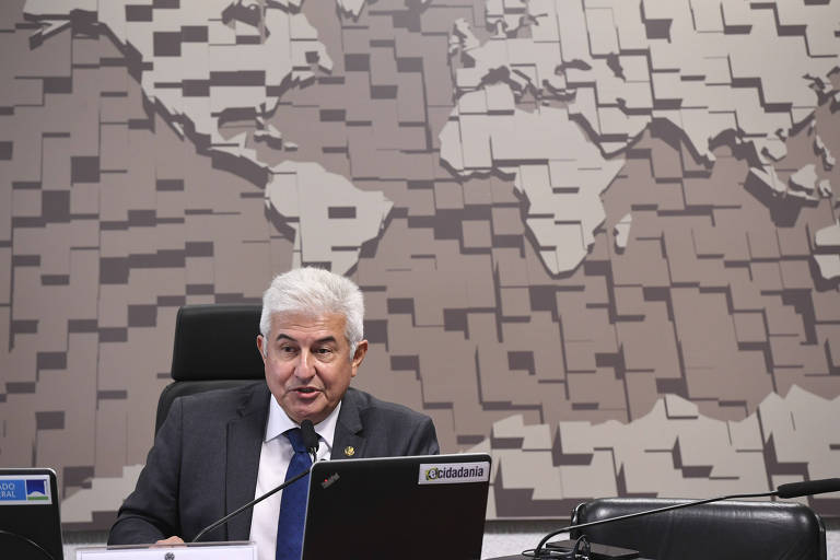 Um homem de cabelo grisalho está sentado em uma cadeira em frente a um laptop， em um ambiente de reunião. Ao fundo， há uma parede com um padrão que representa um mapa-múndi em tons de bege e marrom. O homem usa um terno escuro e uma gravata azul， e parece estar participando de uma videoconferência ou reunião.
