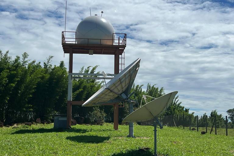 A imagem mostra uma estação de radar com uma cúpula branca no topo de uma estrutura elevada. Na parte inferior， há três antenas parabólicas voltadas para diferentes direções. O fundo é composto por uma área verde com grama e algumas árvores ao longe， sob um céu parcialmente nublado