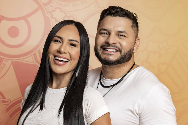 Um casal sorridente posa para a foto. A mulher está à frente, usando uma blusa branca de manga curta e jeans escuros, com cabelo longo e liso. O homem está atrás dela, vestindo uma camiseta branca e segurando a mão dela. Ambos estão sorrindo, com um fundo em tons de laranja e desenhos gráficos.