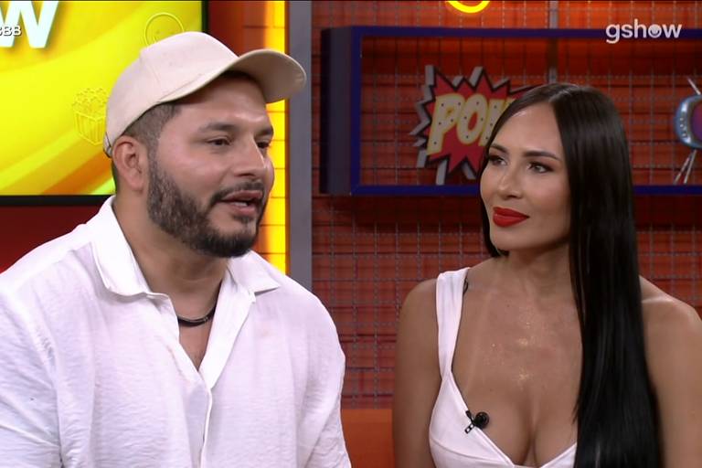 A imagem mostra duas pessoas sentadas em um estúdio de gravação. À esquerda, um homem com barba e cabelo curto, usando uma camisa branca e um boné claro. À direita, uma mulher com cabelo longo e liso, vestindo uma blusa branca e batom vermelho. O fundo é colorido, com elementos gráficos e o logo do programa Bate Papo BBB visível no canto superior esquerdo.