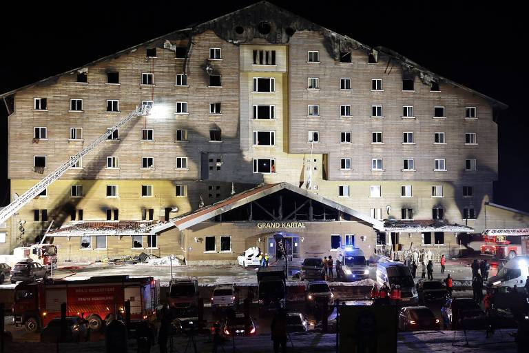 A imagem mostra um hotel chamado 039;Grand Kartal039; em uma noite escura， com evidências de um incêndio. A parte superior do edifício apresenta danos significativos， com janelas quebradas e marcas de fumaça. Veículos de emergência estão estacionados na frente， e bombeiros estão utilizando escadas para combater o fogo. Há uma multidão de pessoas na área， observando a cena.