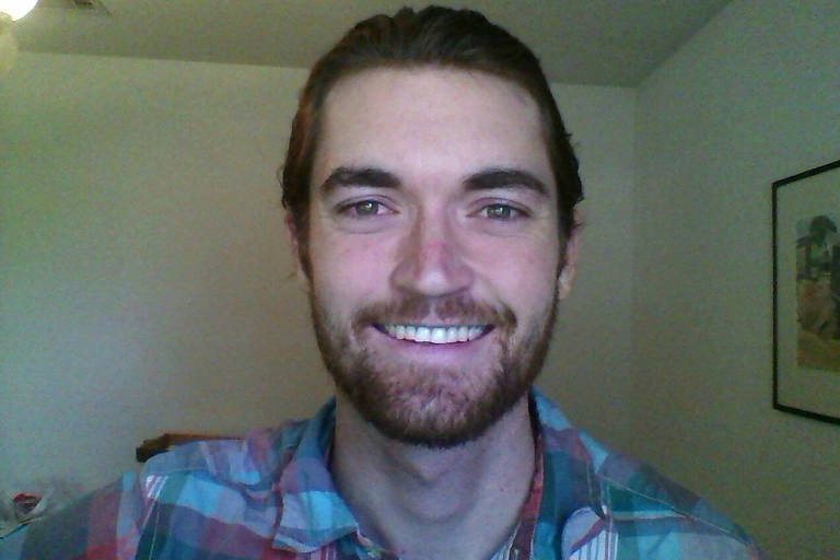 Imagem mostra Ross Ulbricht， um homem com cabelo longo e barba e que está sorrindo para a câmera. Ele usa uma camisa xadrez colorida. Ao fundo， há uma parede clara e um quadro emoldurado. A iluminação é suave.