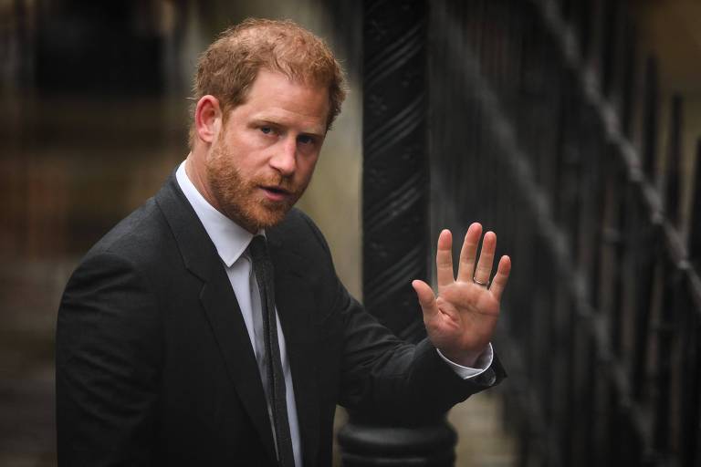 Imagem mostra príncipe Harry， um  homem com cabelo ruivo e barba， vestido com um terno escuro e gravata， e que está acenando com a mão direita. Ele parece estar em um ambiente urbano， com uma grade ao fundo. A expressão facial é séria e ele está olhando para a frente.