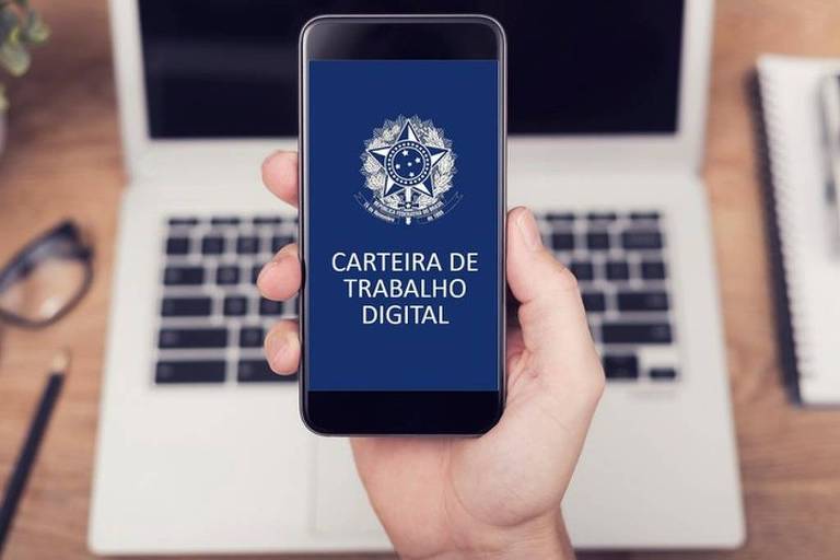 A imagem mostra uma mão segurando um smartphone que exibe a tela da Carteira de Trabalho Digital. O fundo é uma mesa com um laptop e um par de óculos. A tela do smartphone é azul com o texto 039;CARTEIRA DE TRABALHO DIGITAL039; em branco， acompanhado de um símbolo no topo.