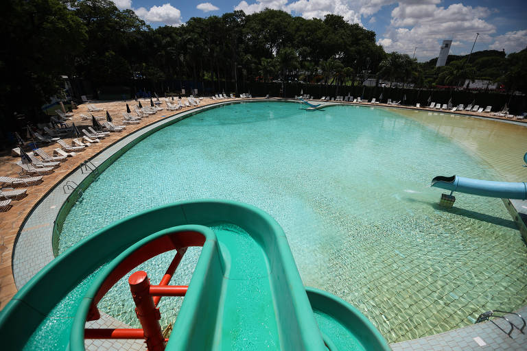 A imagem mostra um parque aquático com uma piscina grande e rasa. No primeiro plano， há um tobogã verde e vermelho que se inclina em direção à água. Ao redor da piscina， há espreguiçadeiras dispostas em fileiras. O céu está claro com algumas nuvens e a vegetação ao fundo é densa.