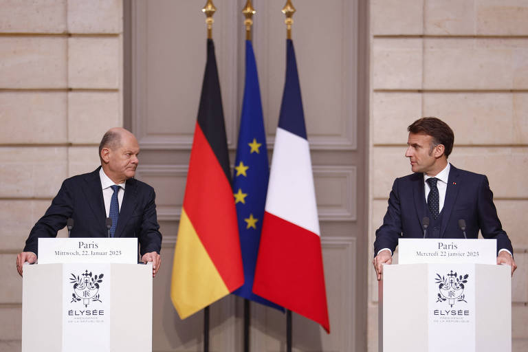 Dois homens de terno e gavatas escuras se olham. À esquerda está Olaf Scholz， premiê alemão， homem branco e calvo. À direita， o presidente francês， Emmanuel Macron， branco de cabelo escuro. Ambos se apoiam em púlpitos brancos com o símbolo do Palácio Eliseu. Ao fundo， as bandeiras da Alemanha， União Europeia e França.