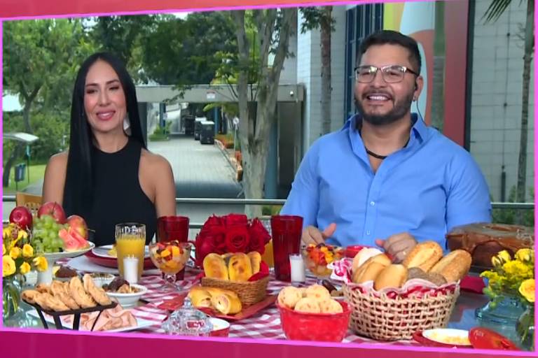 A imagem mostra um cenário de programa de televisão com três apresentadores. À esquerda, uma mulher de cabelo loiro e vestido escuro está em pé, gesticulando. À direita, duas pessoas estão sentadas à mesa, sorrindo. A mesa está cheia de alimentos, incluindo pães, frutas e bebidas. Ao fundo, há uma grande janela com plantas e flores decorativas.