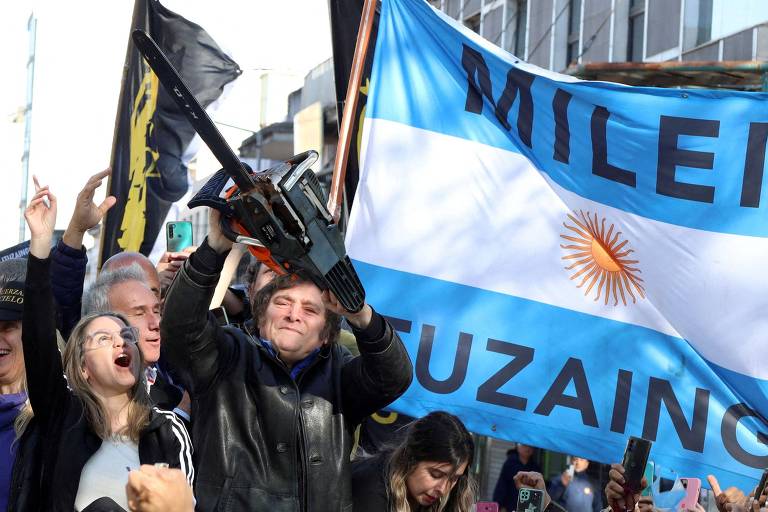 A imagem mostra um grupo de pessoas em uma manifestação. Um homem no centro levanta uma motosserra， enquanto outros ao seu redor estão animados， alguns com as mãos levantadas. Ao fundo， há uma grande bandeira da Argentina， que possui listras horizontais azul e branca， com um sol amarelo no centro. A atmosfera parece ser de celebração ou protesto.