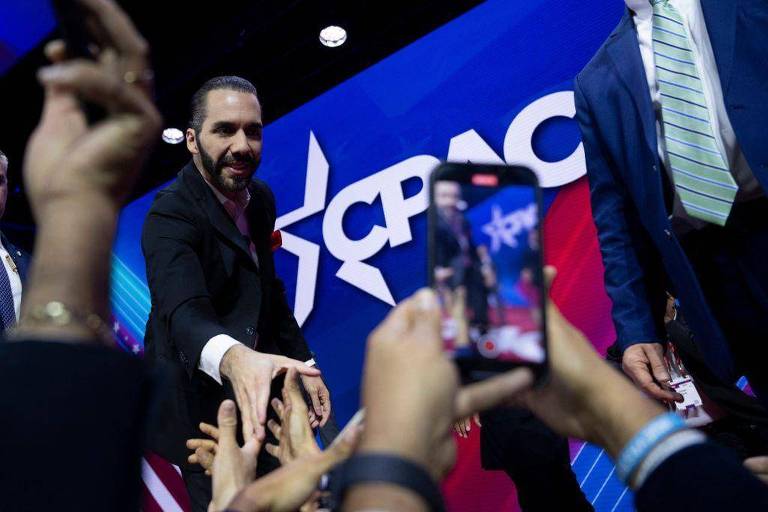 Um homem de terno escuro está cumprimentando pessoas na plateia durante um evento. Ele tem cabelo longo e barba. Ao fundo， há um painel com o logotipo do CPAC em cores azul e branca. Algumas mãos se estendem em direção a ele， enquanto uma pessoa na frente está segurando um celular， filmando a interação.