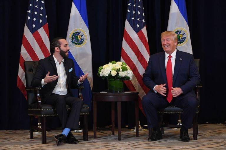 A imagem mostra dois homens sentados em cadeiras em um ambiente formal. À esquerda， um homem com barba e cabelo curto， vestindo um terno escuro e uma camisa branca， gesticula enquanto fala. À direita， um homem com cabelo loiro e uma gravata vermelha， sorrindo. Ao fundo， há bandeiras dos Estados Unidos e de El Salvador， além de um arranjo de flores brancas em uma mesa entre eles.