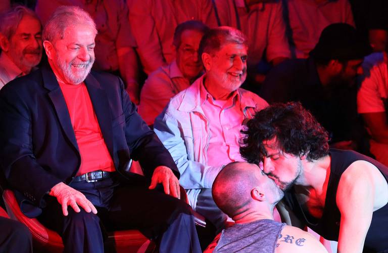 A imagem mostra um grupo de pessoas em um evento, com foco em dois homens se beijando no centro. À esquerda, Lula, sorrindo, vestido com um blazer escuro e uma camiseta vermelha, observa a cena. Ao fundo, outras pessoas assistem, algumas sorrindo. A iluminação é predominantemente em tons de vermelho.