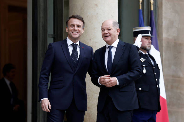 Dois homens em trajes formais estão sorrindo e posando para a câmera na entrada de um edifício. Um deles está à esquerda， com um terno escuro e gravata， enquanto o outro， à direita， também usa um terno escuro. Ao fundo， há um membro da segurança em uniforme， e bandeiras visíveis nas cores da França.