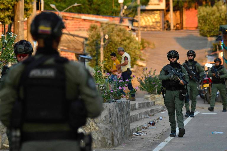 A imagem mostra um grupo de policiais armados em uma rua urbana. Os policiais estão vestidos com uniformes táticos e coletes à prova de balas， alguns deles estão em movimento enquanto outros permanecem em posição. Ao fundo， há casas e vegetação， indicando um ambiente residencial. A iluminação sugere que a cena pode ter sido capturada durante o final da tarde