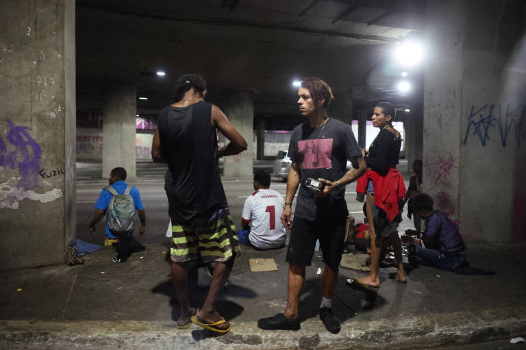 A imagem mostra um grupo de pessoas em uma área coberta. Algumas pessoas estão sentadas no chão, enquanto outras estão em pé. O ambiente é mal iluminado, com luzes artificiais visíveis. As paredes têm grafites