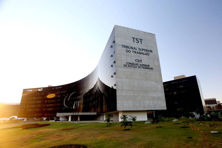 Imagem do edifício do Tribunal Superior do Trabalho (TST) no Brasil， com uma fachada moderna e arquitetonicamente distinta. O prédio é predominantemente de cor clara， com uma parte escura e possui um design curvo. O sol está se pondo ao fundo， iluminando a cena.