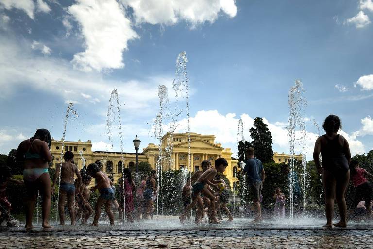 A imagem mostra várias crianças brincando em uma fonte de água， com jatos de água saindo do chão. Ao fundo， há um edifício amarelo e um céu parcialmente nublado. As crianças estão molhadas e parecem se divertir， enquanto a água espirra ao seu redor.