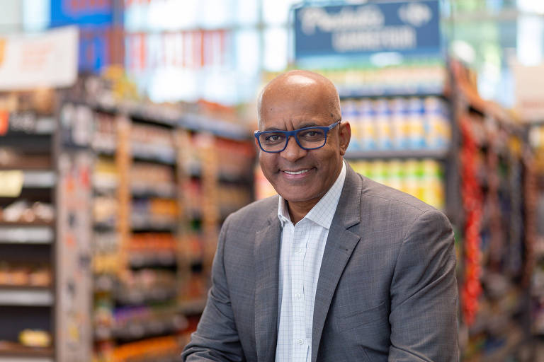 Um homem sorridente está posando em um supermercado. Ele usa um terno cinza e uma camisa branca， além de óculos. Ao fundo， prateleiras com produtos variados estão visíveis， criando um ambiente de loja.