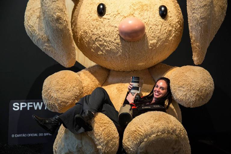 Uma mulher está sentada entre as pernas de um coelho de pelúcia gigante， que é de cor bege claro. Ela segura um objeto em suas mãos e sorri para a câmera. O coelho tem orelhas longas e um nariz rosa. Ao fundo， há uma placa com a inscrição 039;SPFW039;.