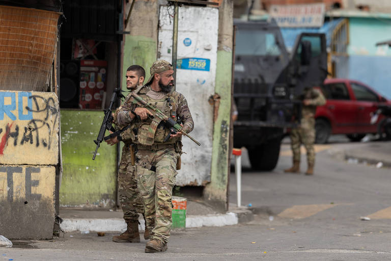 A imagem mostra dois soldados armados em uma rua urbana. Eles estão vestidos com uniformes camuflados e carregam armas. Ao fundo， há um veículo militar e uma parede com grafites. O ambiente parece ser uma área de conflito ou operação policial.
