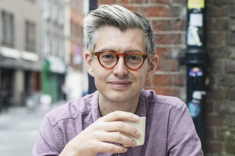 Um homem sentado em uma mesa de madeira em um café ao ar livre. Ele está segurando uma pequena xícara e olhando para a câmera. O homem tem cabelo curto e grisalho， usa óculos com armação laranja e uma camisa de manga longa de cor lilás. Ao lado dele， há uma jarra de vidro vazia sobre a mesa. Ao fundo， é possível ver um ambiente urbano com prédios e algumas pessoas.