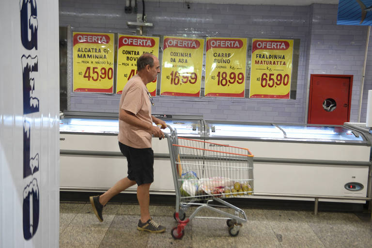 Um homem está caminhando em um supermercado， empurrando um carrinho de compras. Ao fundo， há um balcão de congelados com várias placas de preços em destaque. As placas são amarelas e vermelhas， com preços visíveis. O homem está vestido com uma camisa clara e shorts escuros.
