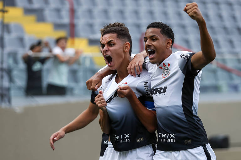 Dois jogadores de futebol estão se abraçando e comemorando em um estádio. Um deles está com os braços levantados e expressa alegria， enquanto o outro sorri e parece estar gritando de felicidade. Ao fundo， há algumas pessoas nas arquibancadas.
