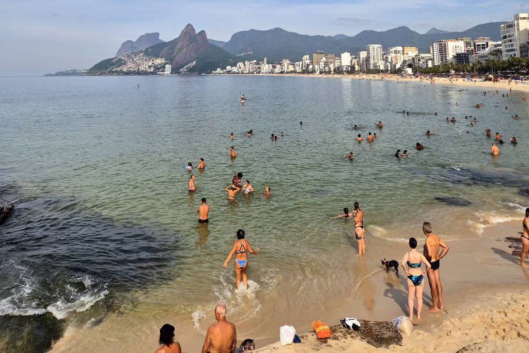  A imagem mostra uma praia com várias pessoas nadando e se divertindo na água. O céu está parcialmente nublado， e ao fundo， é possível ver montanhas e edifícios à beira-mar. A areia é clara e algumas rochas estão visíveis na água.