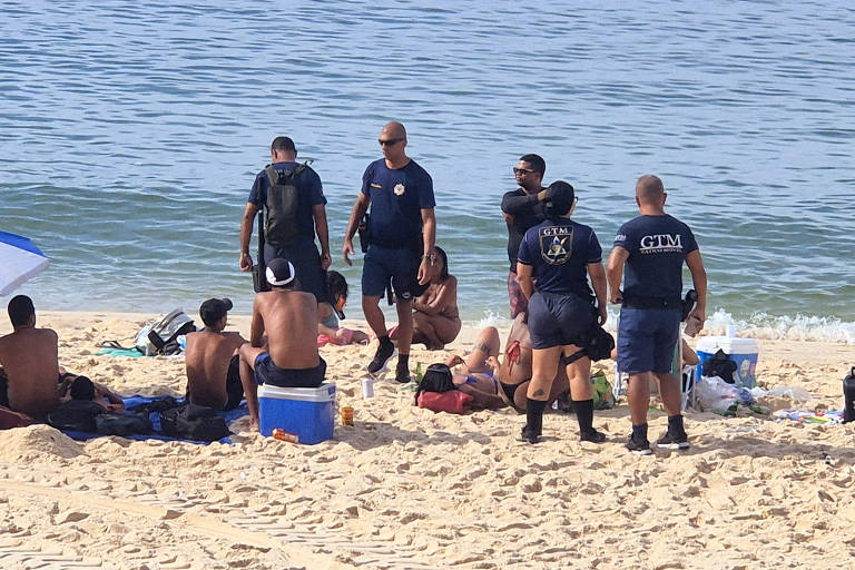 A imagem mostra um grupo de pessoas na praia， incluindo vários homens em trajes de banho e alguns em uniformes de resgate. Eles estão reunidos em uma área de areia próxima ao mar， com algumas pessoas sentadas e outras em pé. Há também objetos como coolers e toalhas espalhados pela areia. O mar está calmo ao fundo.