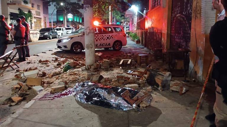 A imagem mostra uma cena noturna em uma rua， onde há um veículo de emergência estacionado. O chão está coberto de detritos， incluindo caixas e outros objetos. Ao fundo， há uma parede com grafite e iluminação de emergência， criando um ambiente tenso. Algumas pessoas estão visíveis， mas não é possível identificar suas ações.