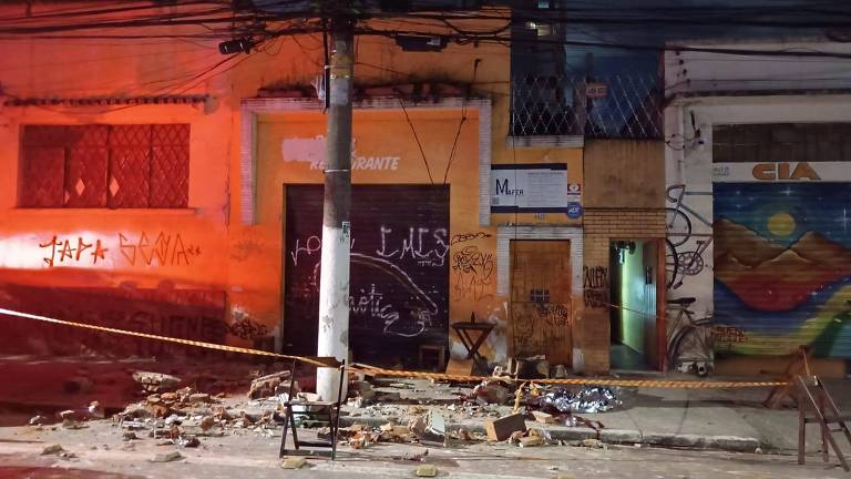 A imagem mostra uma rua à noite com edifícios danificados. À esquerda， há uma parede amarela com janelas escuras e uma porta preta. No centro， um poste de luz está iluminado， e há destroços no chão. À direita， uma parede colorida com grafite é visível， e uma porta verde está entreaberta. A iluminação é predominantemente laranja， sugerindo uma atmosfera de emergência.