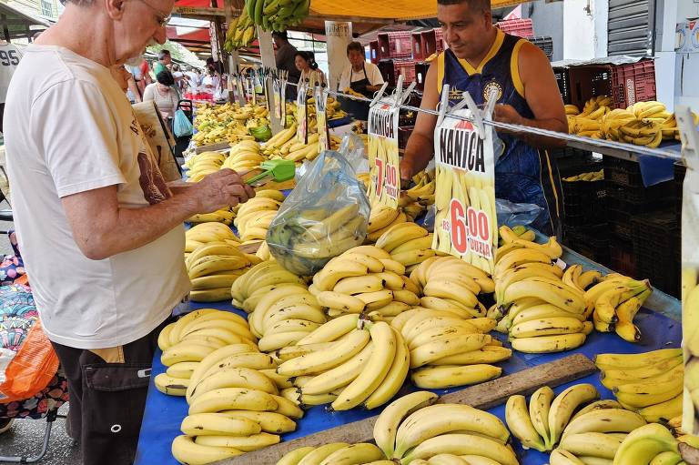 A imagem mostra uma feira de frutas com várias bancadas cheias de bananas. Um homem idoso， vestindo uma camiseta branca， está olhando para seu celular enquanto segura uma sacola plástica. À sua frente， há uma grande quantidade de bananas dispostas em uma mesa azul. Um vendedor， usando uma camiseta sem mangas azul， está pesando bananas em uma balança. Placas indicam os preços das bananas， com valores de R$ 6，00 e R$ 7，00. Ao fundo， outras pessoas estão comprando frutas.