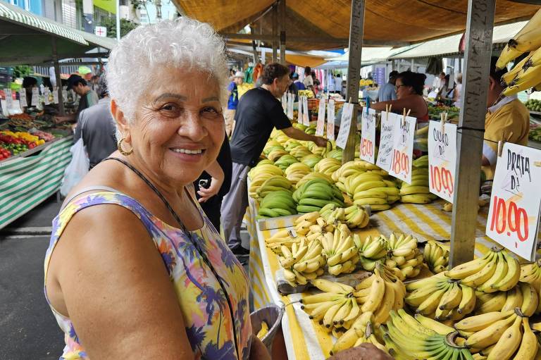 A imagem mostra uma feira de frutas ao ar livre， com barracas cobertas. Em primeiro plano， uma mulher idosa sorri， usando uma blusa colorida. Ao fundo， há várias barracas com frutas， incluindo muitas bananas， e placas de preços visíveis， como 039;Prata R$ 10，00039; e 039;Ouro R$ 10，00039;. Várias pessoas estão comprando e vendendo frutas.