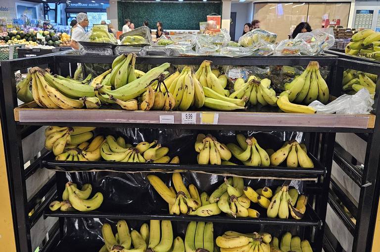 A imagem mostra uma prateleira de supermercado com diferentes tipos de bananas. À esquerda， há um cartaz amarelo com o texto 039;Tipos de bananas039;， e abaixo dele， as descrições 039;Prata: Ideal para doces como a bananada039; e 039;Maçã: Recomendado para bebês e idosos039;. As bananas estão organizadas em três níveis， com algumas maduras e outras ainda verdes. Ao fundo， é possível ver outras frutas e pessoas fazendo compras.