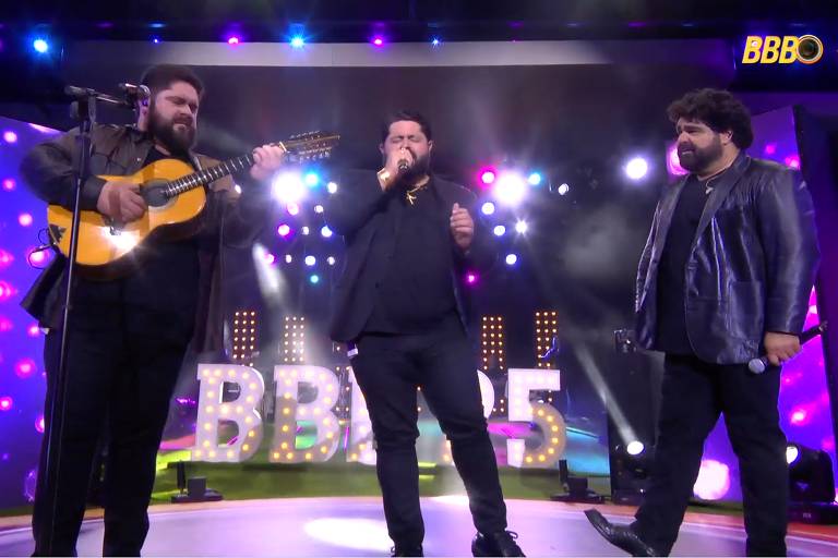 Três homens estão se apresentando em um palco iluminado. O primeiro à esquerda toca um violão e usa uma camisa escura. O segundo, ao centro, canta com um microfone e veste uma camisa preta. O terceiro, à direita, observa e segura um microfone, usando um paletó preto. Ao fundo, há uma iluminação colorida e o letreiro 'BBB 25' em destaque.