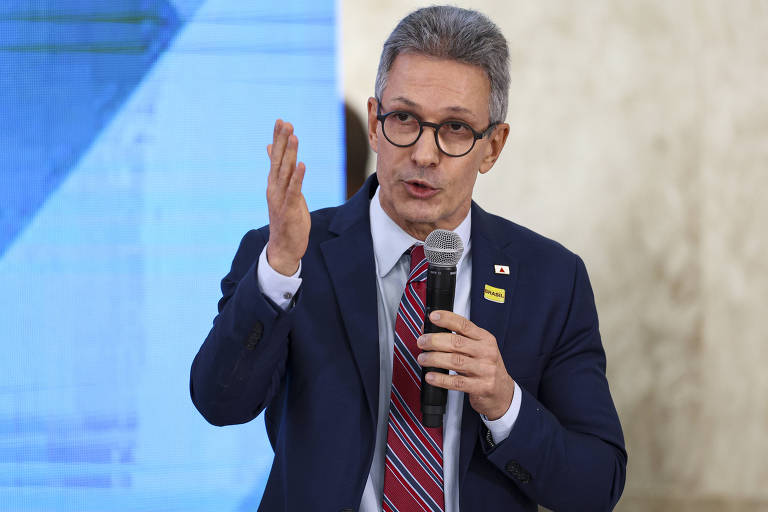 Um homem de óculos， com cabelo grisalho e usando um terno escuro， está falando ao microfone. Ele gesticula com a mão direita enquanto se expressa. Ao fundo， há uma tela com um padrão azul e uma parede clara.