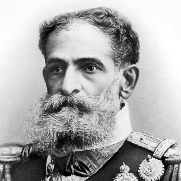 A imagem é um retrato em preto e branco de um homem com uma barba longa e grisalha, usando um uniforme militar adornado com várias medalhas e condecorações. Ele tem uma expressão séria e está olhando para frente. O fundo da imagem é desfocado, destacando o sujeito.