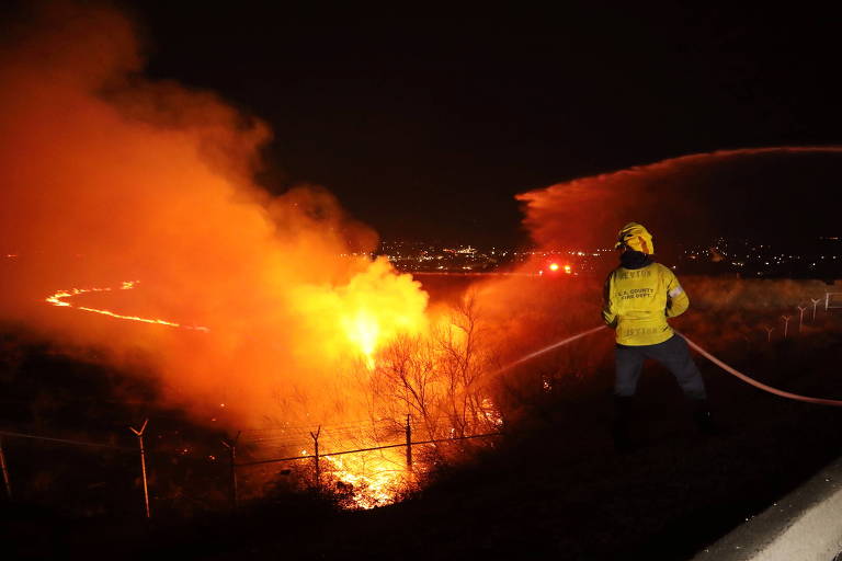 A imagem mostra um bombeiro lutando contra um incêndio à noite. O fogo é intenso， emitindo uma luz laranja brilhante e fumaça densa. O bombeiro está usando um uniforme amarelo e segurando uma mangueira， direcionando água para o fogo. Ao fundo， é possível ver uma cidade iluminada.