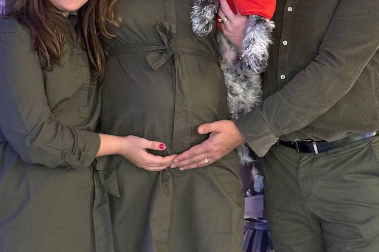 A imagem mostra um casal e uma mulher grávida em um ambiente festivo. A mulher grávida está usando um vestido verde e está com as mãos sobre a barriga. O homem ao lado dela também veste uma camisa verde e está segurando um cachorro pequeno， que usa um chapéu vermelho. A mulher à esquerda também está vestida de verde e sorri para a câmera.