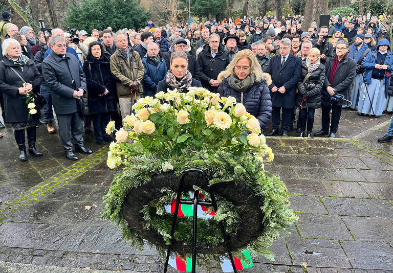 A imagem mostra uma cerimônia de homenagem ao ar livre， com um grupo grande de pessoas reunidas em um espaço público. No primeiro plano， há uma coroa de flores brancas sobre um suporte， com fitas coloridas na base. Ao fundo， várias pessoas estão em pé， algumas com expressões sérias， vestindo roupas formais e escuras， observando a cerimônia. O ambiente é de respeito e solenidade.