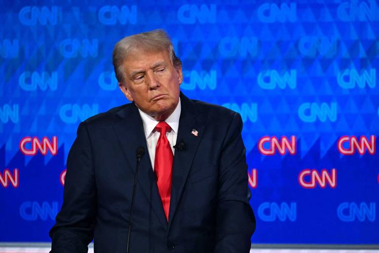 Um homem com cabelo loiro grisalho e uma gravata vermelha está em um palco， com uma expressão pensativa. O fundo é azul com o logotipo da CNN repetido. Ele está vestido com um terno escuro.
