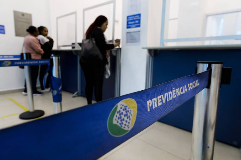 A imagem mostra o interior de uma agência， onde várias pessoas estão em fila para atendimento. No primeiro plano， há uma fita de isolamento azul com o texto 039;PREVIDÊNCIA BRASIL039; e um logotipo. Ao fundo， algumas pessoas estão em pé， aguardando para serem atendidas em um balcão.