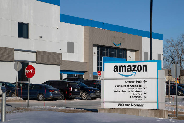 A imagem mostra a fachada de um centro de distribuição da Amazon. À frente， há um grande sinal com o logotipo da Amazon e informações sobre direções. O edifício é de cor clara， com um teto azul e várias janelas. Há carros estacionados na área e um sinal de pare visível.