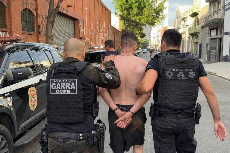 A imagem mostra dois policiais em uniformes pretos， um deles com a palavra 039;GARDA039; e o outro com 039;DAS039;， acompanhando um homem sem camisa que está com as mãos algemadas atrás das costas. O cenário é uma rua urbana， com um veículo policial estacionado ao fundo e edifícios ao lado.