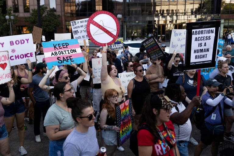 A imagem mostra um grupo de pessoas participando de um protesto. Muitas delas seguram cartazes com mensagens sobre direitos reprodutivos e acesso ao aborto. Há uma placa com um símbolo de proibição. O ambiente é urbano， com prédios ao fundo e algumas pessoas usando óculos escuros e roupas coloridas