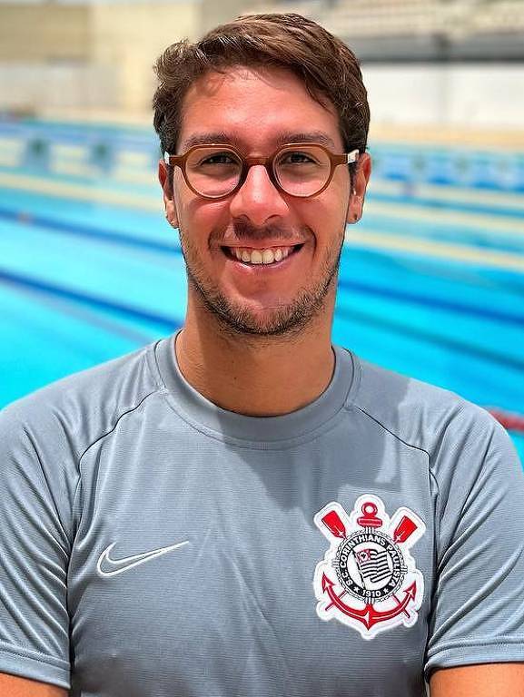 Um homem sorridente está posando em frente a uma piscina olímpica. Ele usa uma camiseta cinza com o logotipo de um clube de futebol. A piscina tem várias raias e está sob um céu nublado. Ao fundo, há arquibancadas vazias.