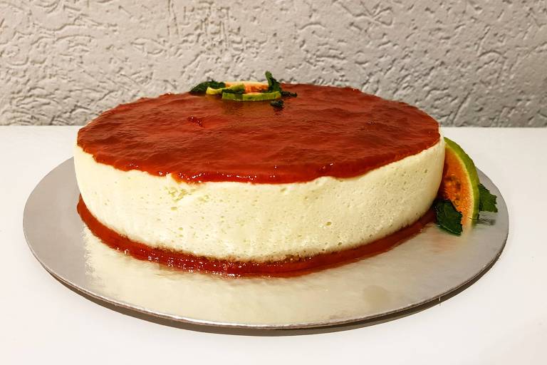 A imagem mostra um cheesecake redondo com uma cobertura vermelha brilhante de goiabada. O cheesecake tem uma base clara e é decorado com uma fatia de fruta e folhas verdes na lateral. O fundo é uma parede texturizada e a superfície onde o cheesecake está colocado é branca.