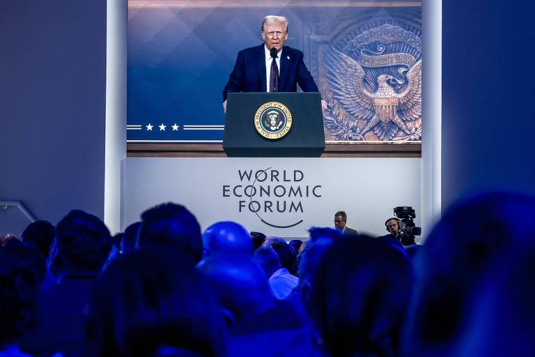 A imagem mostra um palco onde Donald Trump está falando em um evento， com um grande selo presidencial ao fundo. O orador está em frente a um púlpito com o emblema dos Estados Unidos. A plateia é visível， mas as pessoas estão em silhueta devido à iluminação azul predominante.