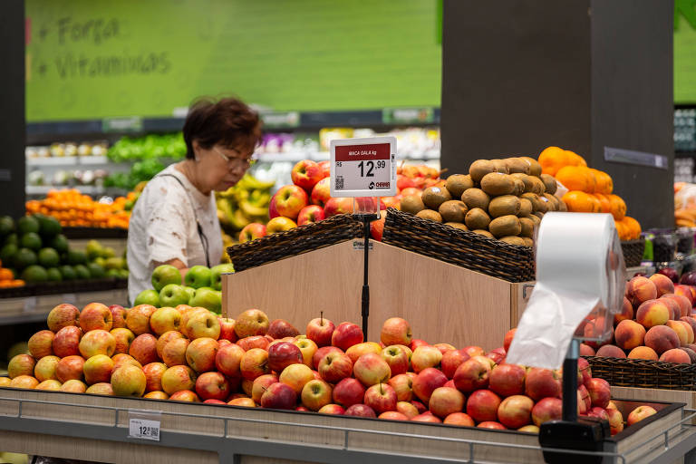 Uma mulher está em um mercado， observando uma seção de frutas. À sua frente， há uma variedade de maçãs em diferentes tons de vermelho e verde， além de batatas e laranjas em uma prateleira. O fundo é verde， com prateleiras de frutas e vegetais visíveis.