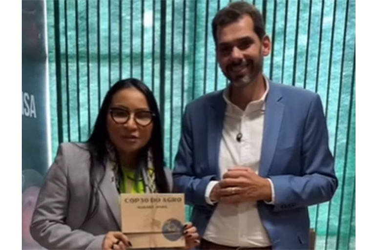 Duas pessoas estão posando para a foto. A mulher à esquerda usa óculos e uma blusa verde sob um paletó cinza， segurando um livro intitulado 039;COP 30 do AGRO039;. O homem à direita está vestido com um paletó azul e uma camisa branca， sorrindo. Ao fundo， há uma parede com listras verticais e uma iluminação suave.