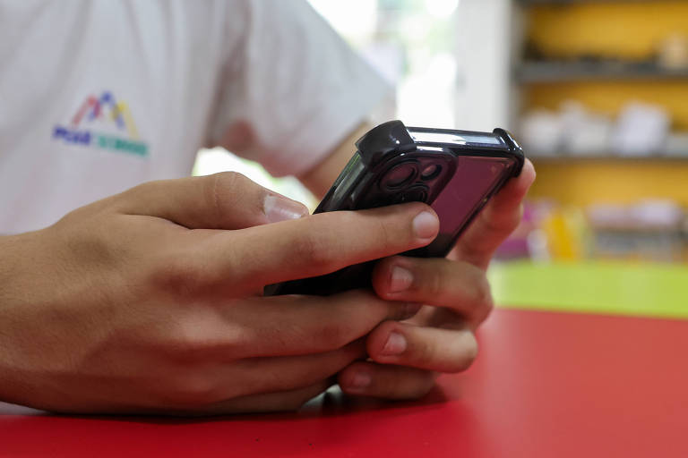 A imagem mostra uma pessoa segurando um celular com as duas mãos. A pessoa está usando uma camiseta branca com um logotipo colorido. O fundo é desfocado， mas é possível ver uma mesa vermelha e prateleiras com objetos coloridos.